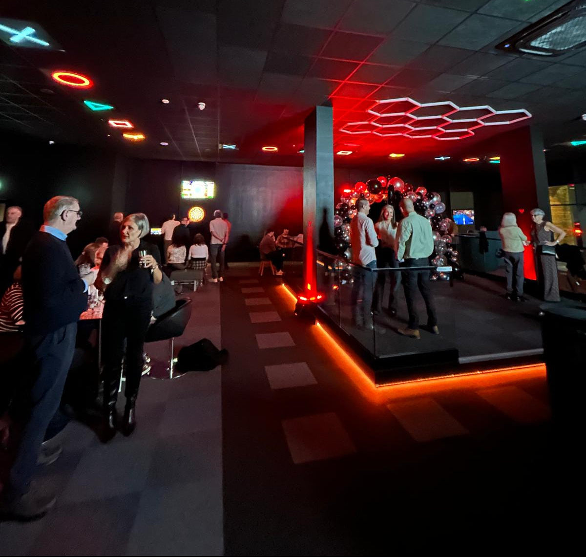 A dimly lit indoor event space or club with multiple people standing and mingling. The room has dark ceiling panels, red and orange lighting accents, and a raised stage area in the background.