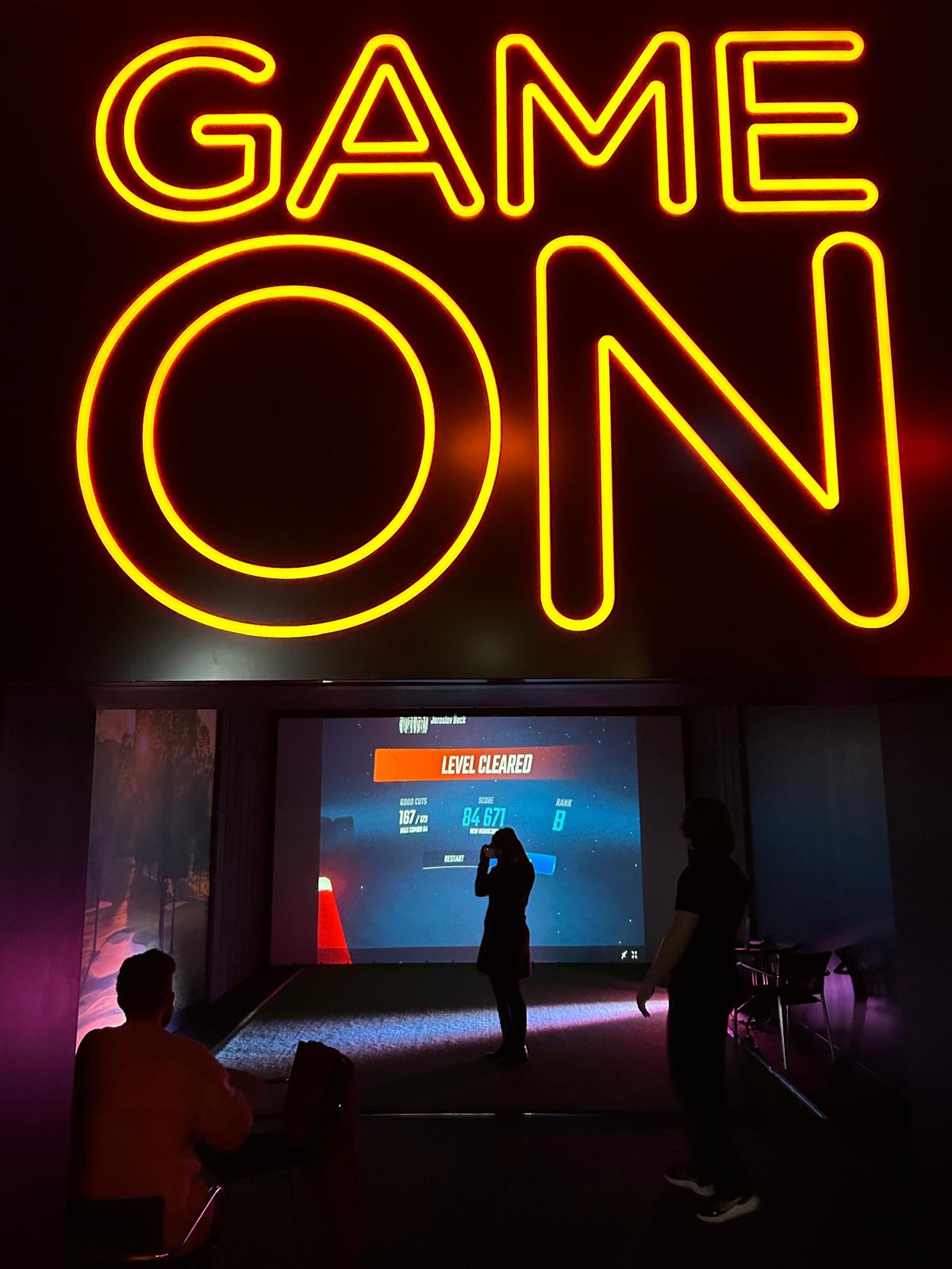 A large glowing yellow neon sign reading 'GAME ON' hangs above a dark gaming setup with a projection screen, silhouetted people, and purple ambient lighting in an indoor event or lounge space.
